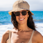 Woman standing in front of the ocean wearing sunglasses, a tan sun hat and a matching tan bikini