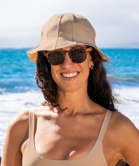Woman standing in front of the ocean wearing sunglasses, a tan sun hat and a matching tan bikini