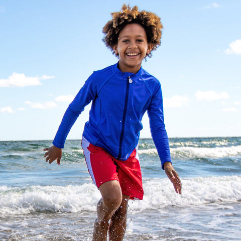 Boy wearing a blue long sleeve rash guard on the beach with red and white swim trunks