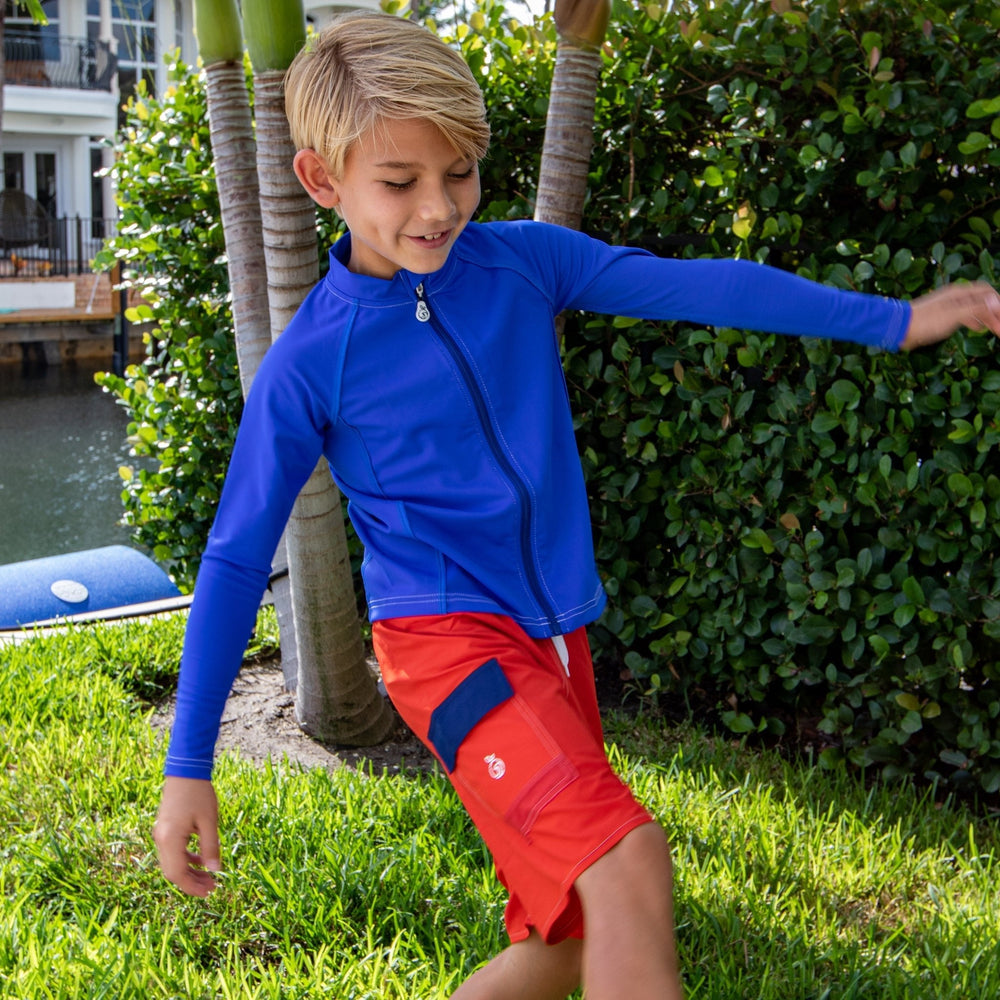 Boy in a rashguard and swim trunks kicking a ball on a lawn