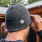 Person wearing a gray knit beanie with a brand logo, outdoors.