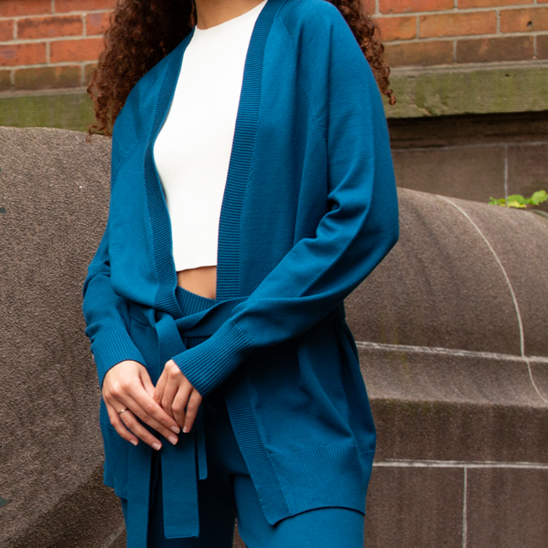 Person wearing a blue cardigan over a white top and dark pants, standing on steps with a brick wall background.